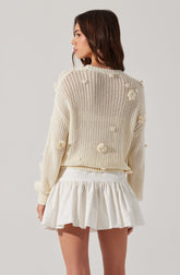 A woman is wearing a cream-colored, oversized knit sweater with textured pom-pom details scattered throughout. She pairs it with a high-waisted, white, ruffled mini skirt.
